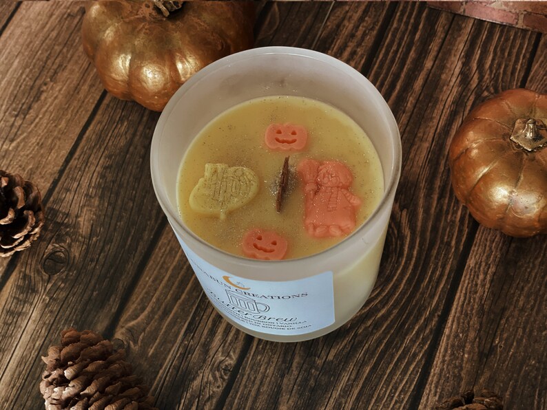May include: A candle in a clear glass jar with a light yellow wax, decorated with small orange pumpkin and figure-shaped wax pieces. The candle sits on a wooden surface with gold pumpkins and pinecones. The label reads "Autumn Creations".