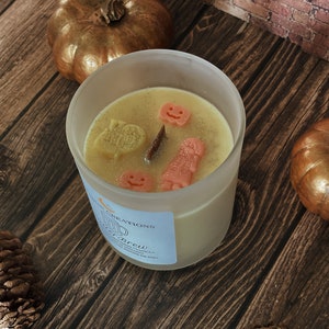 May include: A frosted glass candle with a light yellow wax, decorated with orange and yellow Halloween-themed shapes. The candle sits on a wooden surface with two gold pumpkins and pinecones. The candle has a label that reads "Witch's Brew".