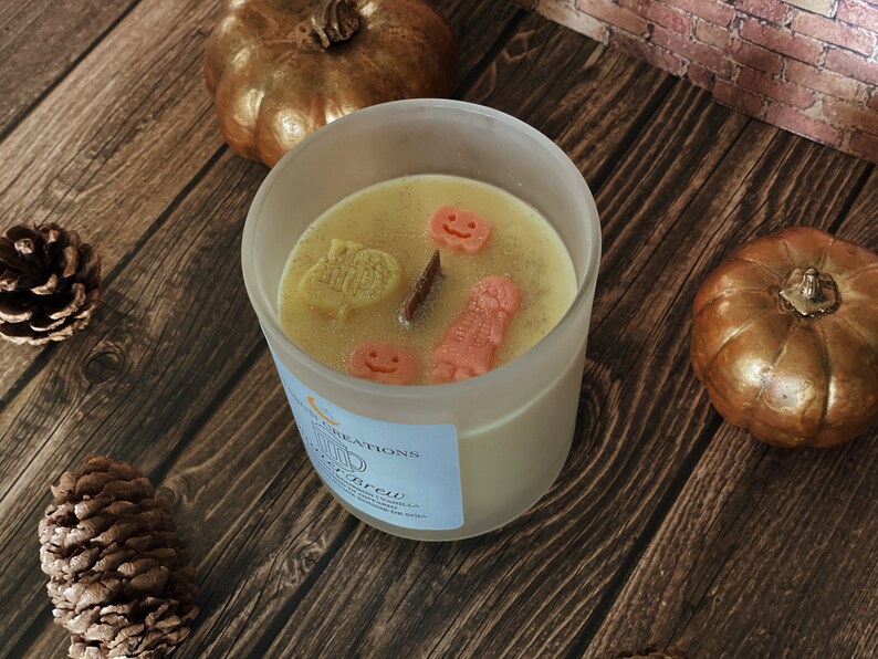 May include: A frosted glass candle with a light yellow wax, decorated with orange and yellow Halloween-themed shapes. The candle sits on a wooden surface with two gold pumpkins and pinecones. The candle has a label that reads "Witch's Brew".