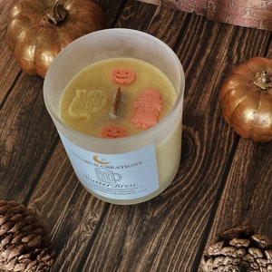 May include: A candle in a clear glass jar with a light yellow wax, decorated with small orange and yellow shapes. The candle is surrounded by two gold pumpkins and pinecones on a wooden surface. The label reads "Butter Brew".