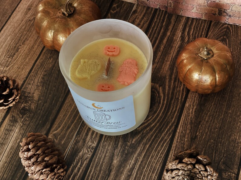 May include: A candle in a clear glass jar with a light yellow wax, decorated with small orange and yellow shapes. The candle is surrounded by two gold pumpkins and pinecones on a wooden surface. The label reads "Butter Brew".