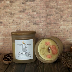 May include: Two glass jar candles with wooden lids. One candle is upright, displaying a label that reads "Butter Brew" and the ingredients. The other candle is on its side, revealing decorative wax shapes. A pinecone and a pumpkin are also visible.