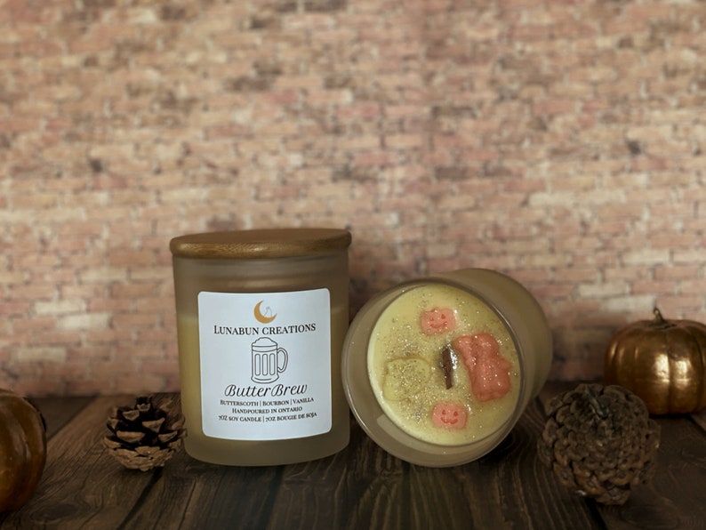May include: Two glass jar candles with wooden lids. One candle is upright, displaying a label that reads "Butter Brew" and the ingredients. The other candle is on its side, revealing decorative wax shapes. A pinecone and a pumpkin are also visible.