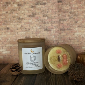May include: A frosted glass candle with a wooden lid, labeled "Butter Brew" with a beer mug graphic. The candle's wax is decorated with small, festive shapes. A pumpkin and pinecone are nearby.
