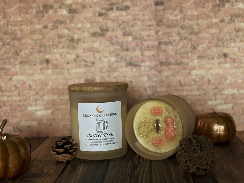 May include: A frosted glass candle with a wooden lid, labeled "Butter Brew" with a beer mug graphic. The candle's wax is decorated with small, festive shapes. A pumpkin and pinecone are nearby.