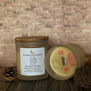 May include: Two frosted glass candles with wooden lids. One candle is upright, the other is tilted, revealing a decorative wax top with pumpkin and ghost shapes. The label reads "Butter Brew" with scent details and "Handpoured in Ontario."