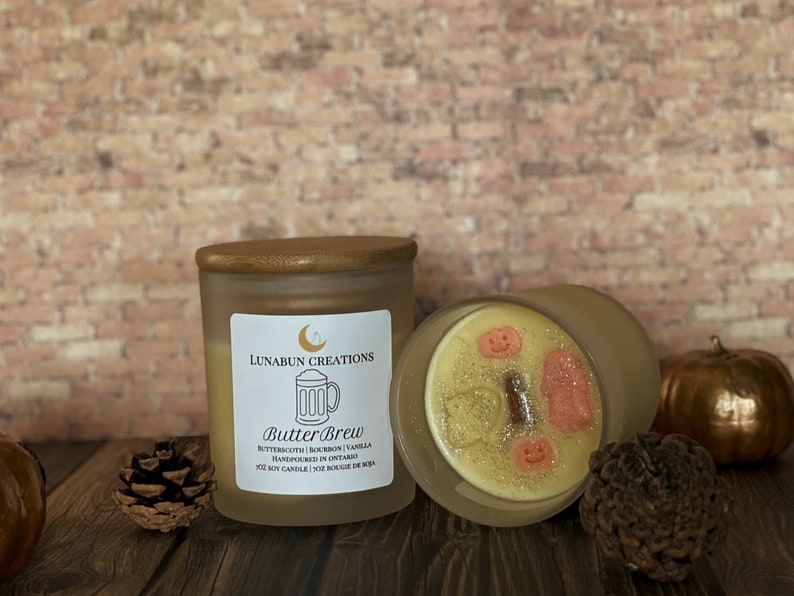 May include: Two frosted glass candles with wooden lids. One candle is upright, the other is tilted, revealing a decorative wax top with pumpkin and ghost shapes. The label reads "Butter Brew" with scent details and "Handpoured in Ontario."