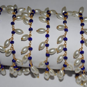 K&ouml;nnte beinhalten: Eine Str&auml;hne aus wei&szlig;en Perlen mit blauen Glasperlen und goldenen Kettengliedern. Die Perlen sind in einem kaskadierenden Muster aufgereiht.