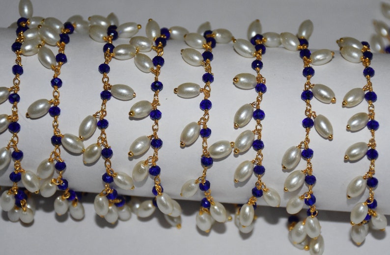 K&ouml;nnte beinhalten: Eine Str&auml;hne aus wei&szlig;en Perlen mit blauen Glasperlen und goldenen Kettengliedern. Die Perlen sind in einem kaskadierenden Muster aufgereiht.