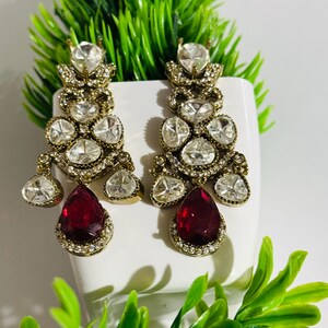 May include: A pair of dangle earrings with a gold tone setting and clear and red stones. The earrings feature a teardrop-shaped red stone at the bottom.