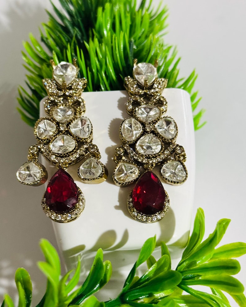 May include: A pair of dangle earrings with a gold tone setting and clear and red stones. The earrings feature a teardrop-shaped red stone at the bottom.