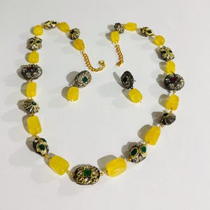 Puede incluir: Un conjunto de collar y pendientes con cuentas de piedras preciosas amarillas y adornos metálicos. El collar y los pendientes tienen una cadena dorada y elementos decorativos con detalles verdes y rojos.