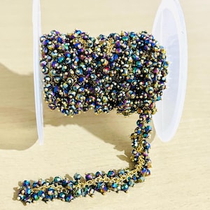 May include: A roll of iridescent beaded chain. The beads are a mix of dark blue, green, and gold, strung on a gold-coloured chain. The chain is partially unraveled, with a portion of the beads hanging down. The roll is white plastic.