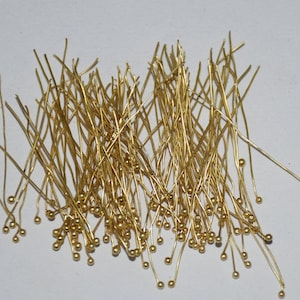 May include: A large pile of gold-colored head pins with round heads. The pins are scattered on a white surface.