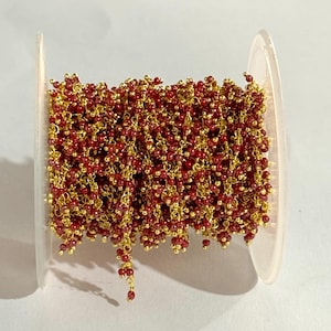 May include: A clear plastic spool holding a beaded chain. The chain is made of small, round, red beads strung on gold-coloured wire. The beads are clustered together, creating a textured effect. The chain is wound around the spool.