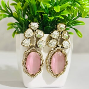 Può includere: Un paio di orecchini pendenti con una pietra ovale rosa circondata da una cornice in metallo dorato e strass trasparenti. La parte superiore degli orecchini presenta un gruppo di strass trasparenti.