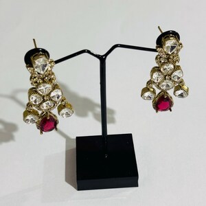 May include: A pair of gold earrings with a teardrop-shaped red stone at the bottom. The earrings are adorned with clear rhinestones and have a dangling design.