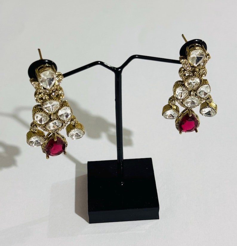 May include: A pair of gold earrings with a teardrop-shaped red stone at the bottom. The earrings are adorned with clear rhinestones and have a dangling design.