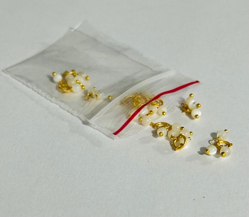 May include: A small plastic bag filled with gold-toned metal charms with white pearl-like beads. The charms are in the shape of a circle with a small loop at the top.