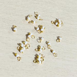 Puede incluir: Una colección de pequeñas piezas de joyería doradas con detalles de perlas blancas. Las piezas están dispersas sobre una superficie de madera clara, y algunas parecen ser pendientes o colgantes.