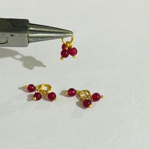 May include: Three gold-toned charms with three red beads each. The charms are designed to be attached to a necklace or bracelet.