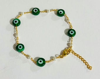 May include: Gold-toned chain bracelet featuring green evil eye beads and small white pearls. The bracelet has an adjustable chain closure. The evil eye beads are a deep green with a white and black center.