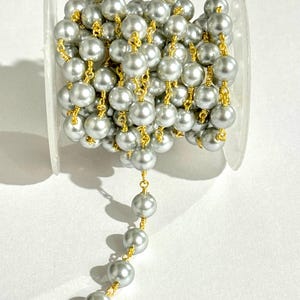 May include: A spool of gray pearl beads strung on gold-colored wire. The pearls are round and evenly sized, creating a continuous strand. The beads are wound around a clear plastic spool, ready for use in jewelry making or craft projects.