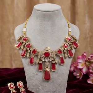 May include: A gold-toned necklace and matching earrings set. The necklace features large, rectangular, and square red gemstones, with smaller clear stones. The earrings have a similar design. The jewelry is displayed on a gray mannequin head.