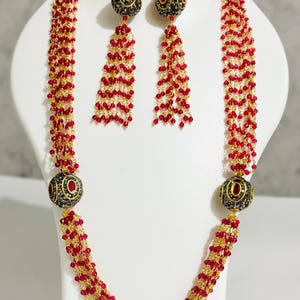 May include: A red beaded necklace and earring set. The necklace features multiple strands of red beads with gold-coloured accents and decorative black and gold spheres. The earrings have a similar design, with red bead tassels.