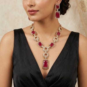 May include: A gold-toned necklace and earring set featuring large, rectangular, ruby-red gemstones. The necklace has a pendant with a large ruby, and the earrings have matching ruby stones. The jewelry is set with small, clear accent stones.