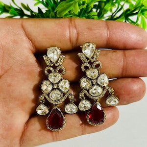 May include: A pair of gold dangle earrings featuring red and clear stones. The earrings are teardrop shaped and decorated with numerous smaller stones.
