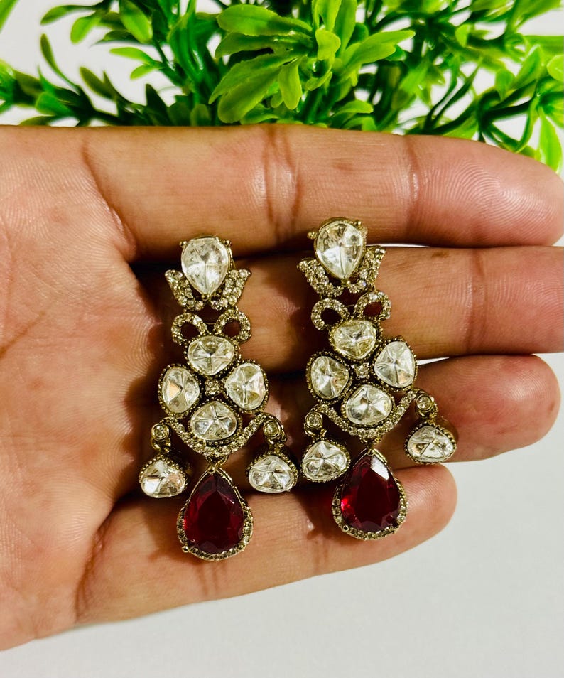 May include: A pair of gold dangle earrings with red and clear stones. The earrings have a teardrop shape and are adorned with multiple smaller stones.