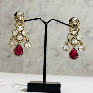 May include: A pair of dangle earrings with a red teardrop-shaped stone at the bottom. The earrings are made of gold and have a cluster of clear stones.