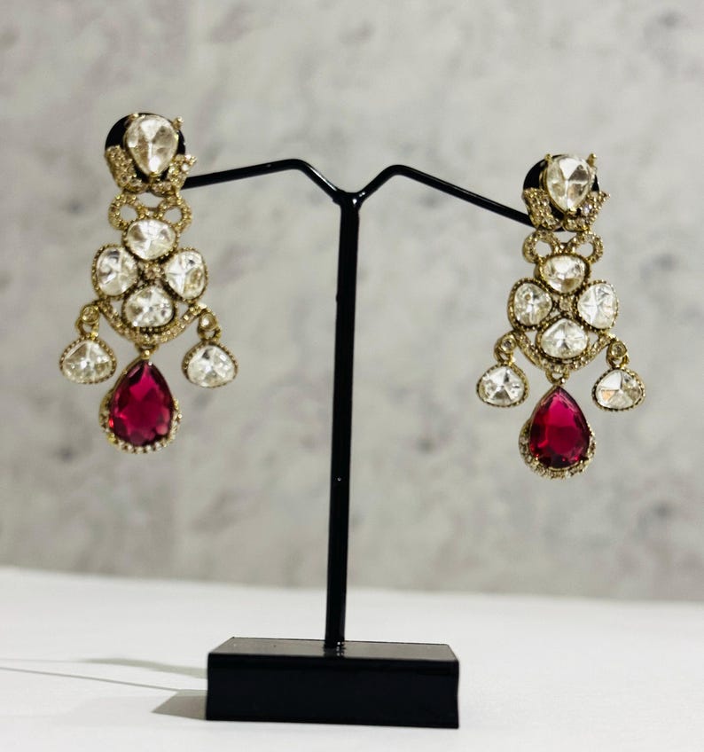 May include: A pair of dangle earrings with a red teardrop-shaped stone at the bottom. The earrings are made of gold and have a cluster of clear stones.