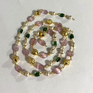以下が含まれることがあります： ピンク、緑、白のビーズが混ざり合ったロングネックレス。金色のテクスチャードアクセントが散りばめられています。ネックレスは、楕円形、丸型、ファセットカットなど、さまざまな形のビーズで構成され、スパイラルパターンに配置されています。
