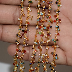 Puede incluir: Primer plano de una mano sosteniendo varios hilos de cadena dorada con cuentas de colores. Las cuentas son pequeñas y de colores variados, incluyendo rojo, azul, verde y blanco.