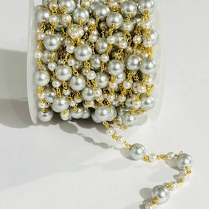 May include: A spool of pearl chain featuring alternating sizes of gray and white faux pearls. The pearls are strung on a gold-colored chain. The chain is partially unraveled, showing the detail of the pearl strand.