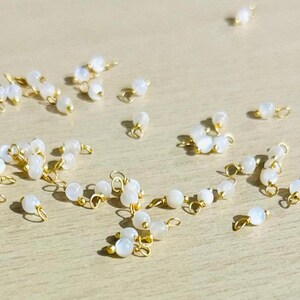 Puede incluir: Una colección de cuentas de piedra lunar blanca con anillos de salto de oro. Las cuentas son redondas y tienen un brillo nacarado. Los anillos de salto son pequeños y delicados.