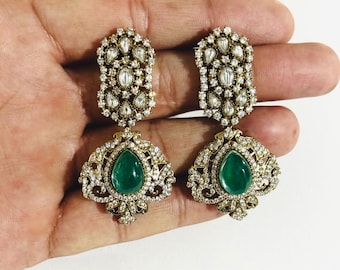 Può includere: Un paio di orecchini pendenti color oro. Ogni orecchino presenta una pietra verde smeraldo a forma di goccia nella parte inferiore, circondata da un design a filigrana e piccole pietre preziose trasparenti. La sezione superiore ha un design simile con più pietre preziose trasparenti.