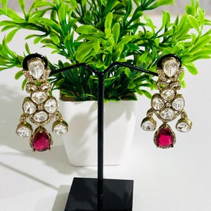 May include: A pair of dangle earrings with a gold tone finish. The earrings feature a cluster of clear rhinestones and a single teardrop-shaped red gemstone.