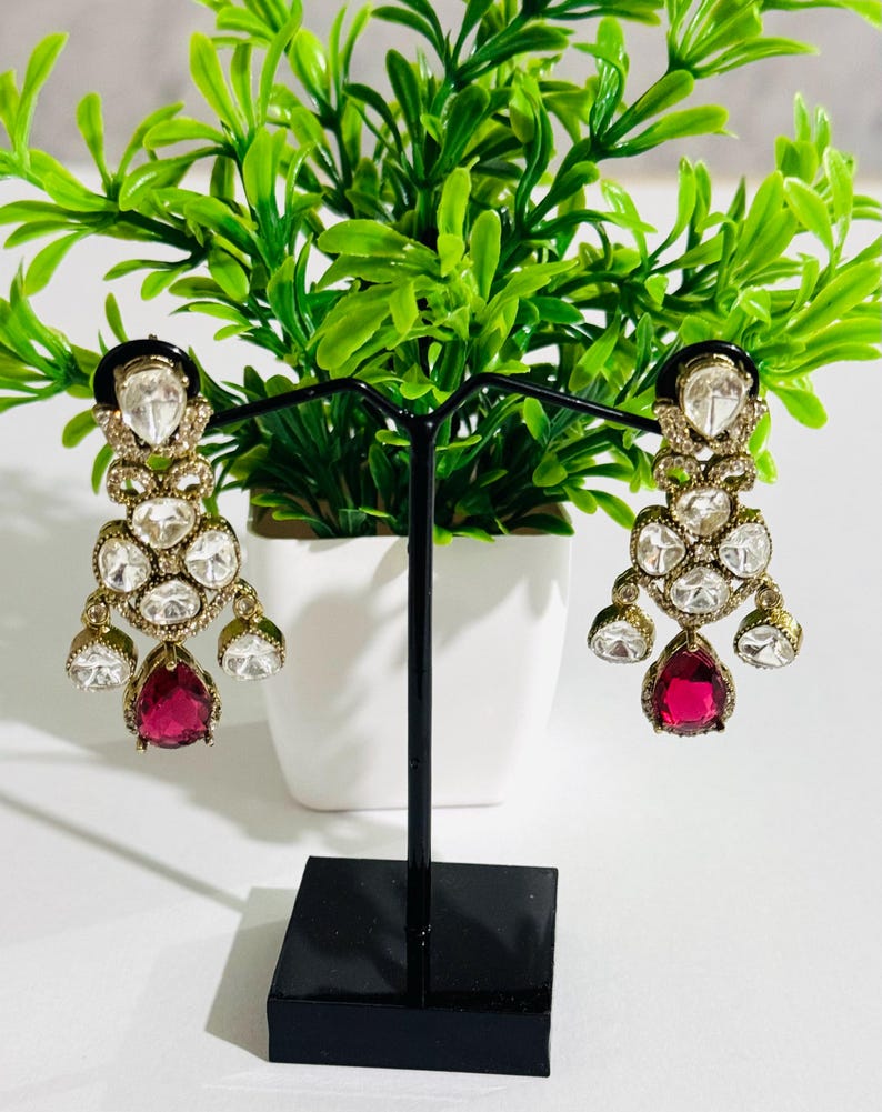 May include: A pair of dangle earrings with a gold tone finish. The earrings feature a cluster of clear rhinestones and a single teardrop-shaped red gemstone.