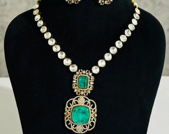 May include: A gold-toned necklace and earring set displayed on a black mannequin. The necklace features a large, square, emerald-green pendant with a decorative gold filigree design. The earrings and necklace are adorned with clear, round stones.