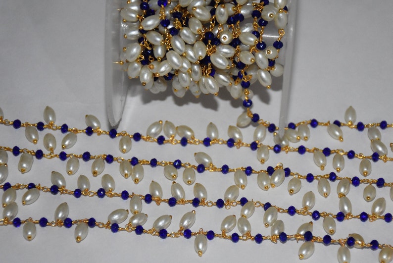 K&ouml;nnte beinhalten: Eine goldene Kette mit wei&szlig;en ovalen Perlen und dunkelblauen Glasperlen. Die Kette ist auf einer Spule aufgewickelt.