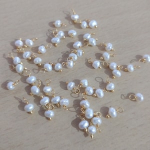 Könnte beinhalten: Eine Sammlung von weißen Perlenanhängern mit goldenen Sprungringen. Die Perlen sind in verschiedenen Größen und Formen. Die Anhänger sind perfekt für die Schmuckherstellung.