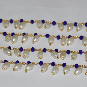 K&ouml;nnte beinhalten: Eine Str&auml;hne aus wei&szlig;en Perlen mit dazwischenliegenden blauen Glasperlen, auf einer goldenen Kette aufgef&auml;delt. Die Perlen sind in einem kaskadierenden Muster angeordnet, was einen zarten und eleganten Look erzeugt.