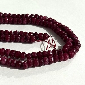 May include: A strand of deep burgundy, faceted round beads arranged in a curved line. The beads have a reflective, polished surface and are strung together on a thin, matching dark red thread, showing a consistent shape and size throughout.