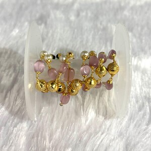 以下が含まれることがあります： 金色のテクスチャードビーズ、ピンクの楕円形のビーズ、白い真珠ビーズが交互に配置された、ビーズジュエリーチェーンのスプール。 チェーンは、白いふわふわの背景に対して、透明なプラスチック製のスプールに表示されています。