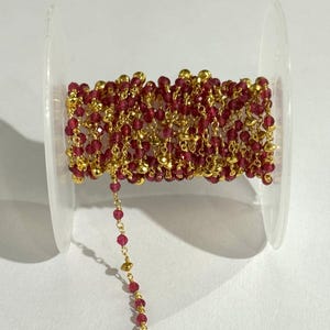 Puede incluir: Un carrete de cadena de cuentas con pequeñas cuentas facetadas de color rojo rubí y detalles metálicos dorados. La cadena está enrollada alrededor de un carrete de plástico transparente, con una parte de la cadena suelta. Suministro de joyería para collares y pulseras.