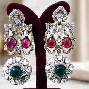 May include: Ornate silver-toned earrings with a floral design. Each earring features a teardrop-shaped red gemstone and a round green gemstone. The earrings are displayed on a burgundy velvet stand.