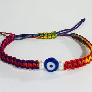 May include: A macrame bracelet with a blue evil eye bead and two white beads. The bracelet features a rainbow of colors, including red, yellow, green, and purple. The bracelet is adjustable.